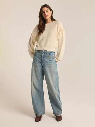 cream crew neck sweater with long sleeves and fringe detail at the shoulders worn with baggy light washed denim jeans 